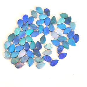 May include: A collection of numerous teardrop-shaped opals in various shades of blue, ranging from light sky blue to deep navy. Each gemstone displays a smooth, polished surface with iridescent flashes of colour throughout the stones.