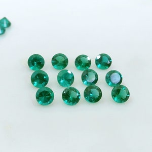 May include: A collection of round, faceted emerald green gemstones. The gemstones are scattered on a white surface, with a few stones grouped together in the upper left corner. The gems have a brilliant cut.