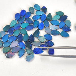 May include: A collection of loose, teardrop-shaped blue opals with iridescent flashes of colour. The stones exhibit various shades of deep blue, cyan, and teal. A pair of silver-toned metal tweezers holds one individual blue opal gemstone.