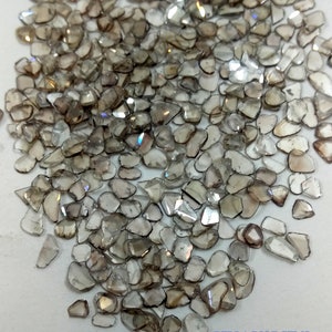 May include: A pile of small, brown, and clear, rough-cut diamonds. The diamonds are scattered on a white surface. The text "BELLAGOLDGEMS" is visible in the lower right corner of the image.