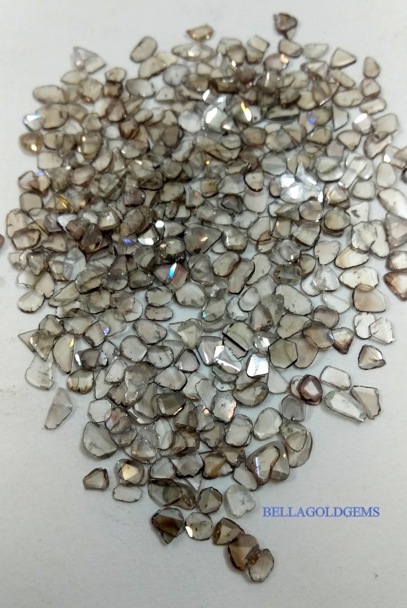 May include: A pile of small, brown, and clear, rough-cut diamonds. The diamonds are scattered on a white surface. The text "BELLAGOLDGEMS" is visible in the lower right corner of the image.