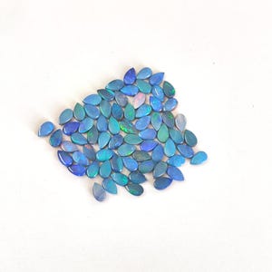 May include: A collection of numerous small, teardrop-shaped opals. Each gemstone displays a vibrant blue base colour with iridescent flashes of green and occasional hints of pink or purple, creating a shimmering effect across the smooth surfaces.