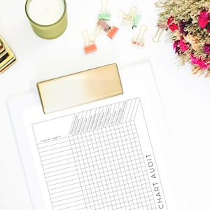 May include: A white clipboard with a gold clip, labeled "CHART AUDIT" with a grid for tracking client and chart IDs. The clipboard is on a white surface with a gold stapler, a candle, and colorful paper clips.