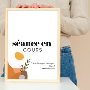 Könnte beinhalten: Ein gerahmtes Schild mit dem Text "séance en cours" in schwarzen Buchstaben auf weißem Hintergrund. Das Schild enthält auch eine kleine Illustration einer Sonne, einer Pflanze und eines Topfes in den Farben Orange, Gelb und Braun. Der Text "Prière de ne pas déranger. Merci!" ist in Schwarz unter der Illustration geschrieben.