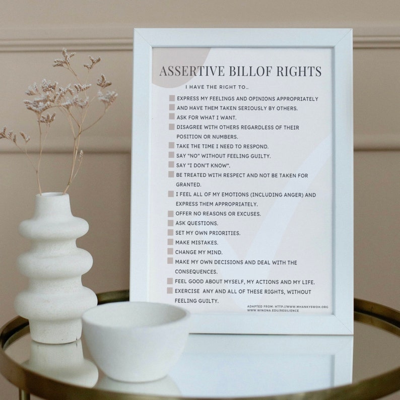 May include: A white framed print with the title "Assertive Bill of Rights" and a list of 14 rights, including expressing feelings, asking for what you want, and setting priorities. The print is on a table with a white vase and a white bowl.