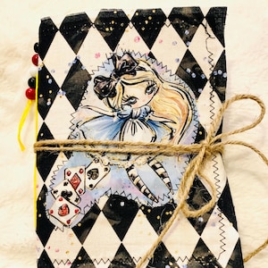 Op de afbeelding: Een handgemaakt tijdschrift met een zwart-wit ruitpatroon en een illustratie van een meisje in een blauwe jurk. Het tijdschrift is vastgebonden met touw en heeft een kralenrug.