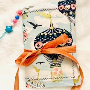 May include: A fabric-covered journal with a hot air balloon design in shades of blue, pink, and gold, tied with an orange ribbon. A beaded charm with a star and flower accents is attached. The journal is approximately 4 inches wide and 6 inches tall.