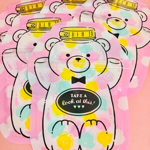 May include: A set of five pink reusable snack bags with a white bear design. The bear is wearing a black bow tie and has a jar lid on its head. The bags have a white background with colorful polka dots and the text "TAKE A look at this!" in a black oval.