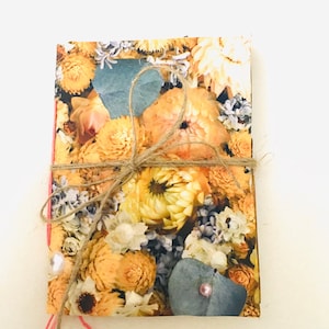 May include: A floral-patterned notebook with a jute twine bow. The cover features a collage of yellow, orange, and white flowers with blue-green leaves. A pink thread and a pearl accent the side of the notebook.