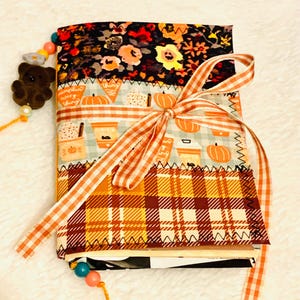 May include: A fabric-covered journal featuring three horizontal panels: a black floral print, a light blue pumpkin and coffee cup pattern, and a yellow and brown plaid section. An orange and white gingham ribbon is tied around the center.
