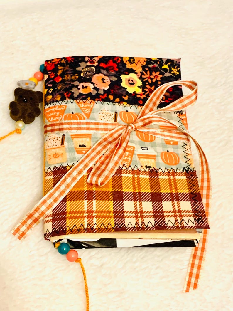 Peut inclure: Un journal recouvert de tissu avec trois panneaux horizontaux : un imprim&eacute; floral noir, un motif bleu clair de citrouilles et de tasses &agrave; caf&eacute;, et une section &agrave; carreaux jaunes et marron. Un ruban vichy orange et blanc est nou&eacute; au centre.