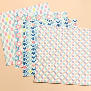 May include: Four sheets of patterned paper with pastel designs. The patterns include arrows, hexagons, waves, and interlocking circles. The colour palette features pink, blue, yellow, and purple on a cream background. The papers are arranged in a layered fan shape.