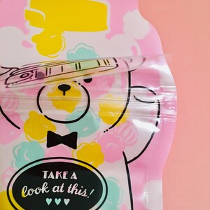 May include: A pink and white reusable snack bag with a cartoon bear design. The bag has a zipper closure and says "TAKE A look at this!" in black text on a black circle.