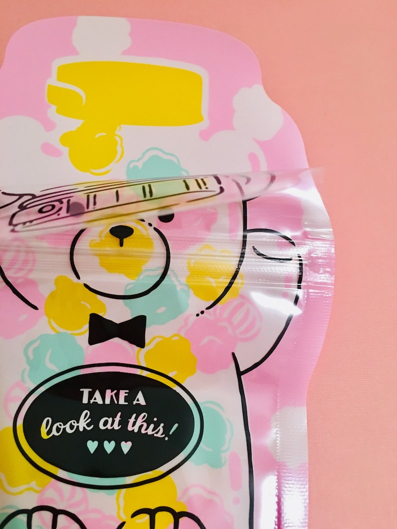 May include: A pink and white reusable snack bag with a cartoon bear design. The bag has a zipper closure and says "TAKE A look at this!" in black text on a black circle.