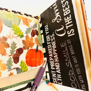 Peut inclure: Un album de scrapbooking ouvert avec une page illustr&eacute;e de citrouilles orange, de glands bruns et de feuilles d'automne color&eacute;es. La page oppos&eacute;e est noire avec le texte blanc : "SENT PREPARED FOR YOU. A SMILE IS THE BEST."
