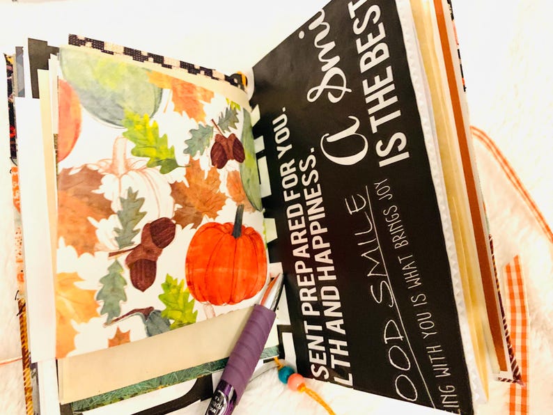 Peut inclure: Un album de scrapbooking ouvert avec une page illustr&eacute;e de citrouilles orange, de glands bruns et de feuilles d'automne color&eacute;es. La page oppos&eacute;e est noire avec le texte blanc : "SENT PREPARED FOR YOU. A SMILE IS THE BEST."
