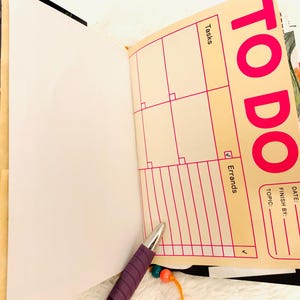 Peut inclure: Un agenda ouvert avec une page couleur cr&egrave;me pr&eacute;sentant un en-t&ecirc;te TO DO rose vif, une grille pour les t&acirc;ches et une section pour les courses. La page inclut des champs pour la date, l'&eacute;ch&eacute;ance et le sujet. Un stylo violet avec une breloque perl&eacute;e repose sur la page.