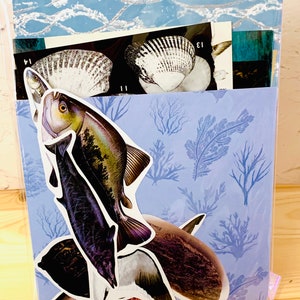 May include: A blue and white patterned paper with a variety of sea-themed stickers, including fish, seashells, and coral. The stickers are arranged in a collage-like fashion.