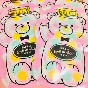 May include: Four pink reusable snack bags with a bear design. Each bag has a white bear with a black bow tie and a jar of colorful candies on its head. The bags have the text "Take a look at this!" printed on them.