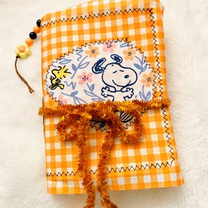 Puede incluir: Bolsa de tela a cuadros naranja y blanco con un diseño de Snoopy y Woodstock. La bolsa está decorada con un parche floral, un lazo naranja decorativo y un cordón de cuentas. Los bordes están cosidos con hilo negro.