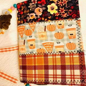 Peut inclure: Un journal recouvert de tissu avec trois panneaux horizontaux : un imprim&eacute; floral noir, une section bleu clair avec des motifs de citrouilles et de tasses de caf&eacute; marqu&eacute;s "pumpkin every thing", et une section inf&eacute;rieure &agrave; carreaux orange et bordeaux.