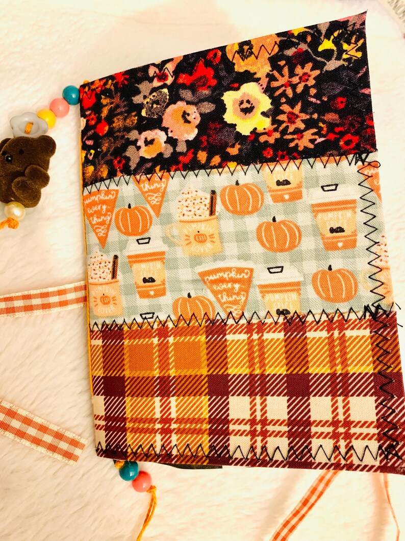 Peut inclure: Un journal recouvert de tissu avec trois panneaux horizontaux : un imprim&eacute; floral noir, une section bleu clair avec des motifs de citrouilles et de tasses de caf&eacute; marqu&eacute;s "pumpkin every thing", et une section inf&eacute;rieure &agrave; carreaux orange et bordeaux.