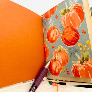 Peut inclure: Un journal ouvert pr&eacute;sentant une page orange unie sur la gauche et une page avec un motif de citrouilles &agrave; l'aquarelle sur la droite. Le motif affiche des citrouilles orange avec des tiges vertes sur un fond bleu sourd. Un stylo violet repose sur les pages.