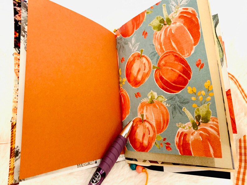 Peut inclure: Un journal ouvert pr&eacute;sentant une page orange unie sur la gauche et une page avec un motif de citrouilles &agrave; l'aquarelle sur la droite. Le motif affiche des citrouilles orange avec des tiges vertes sur un fond bleu sourd. Un stylo violet repose sur les pages.