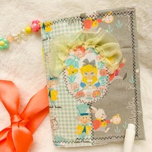 May include: A handmade Alice in Wonderland themed journal. The journal features a patchwork design with illustrations of Alice, the Mad Hatter, and the White Rabbit. It has a yellow ribbon and a beaded charm. The cover includes the word "HATT".