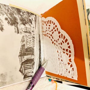 Peut inclure: Un album ouvert pr&eacute;sentant une illustration architecturale vintage en noir et blanc sur la page de gauche et une page orange unie avec un napperon en papier blanc sur la droite. Un stylo &agrave; bille violet repose en diagonale sur les pages.