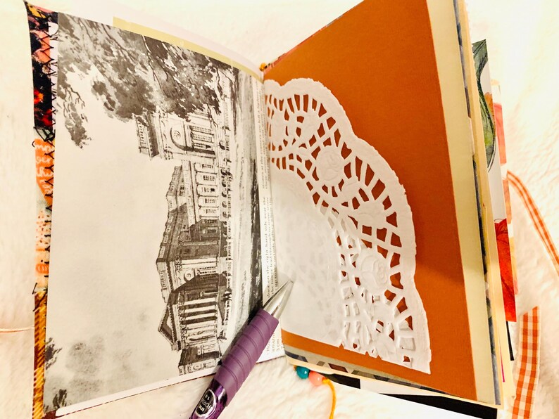 Peut inclure: Un album ouvert pr&eacute;sentant une illustration architecturale vintage en noir et blanc sur la page de gauche et une page orange unie avec un napperon en papier blanc sur la droite. Un stylo &agrave; bille violet repose en diagonale sur les pages.