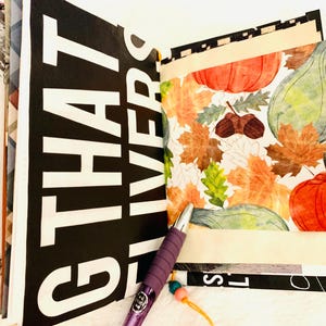 Peut inclure: Un album ouvert pr&eacute;sentant une page avec un grand texte blanc sur fond noir indiquant "THAT LIVERS" et une page adjacente d&eacute;cor&eacute;e d'un motif automnal color&eacute; de citrouilles, de courges et de feuilles d'automne. Un stylo violet est pos&eacute;.