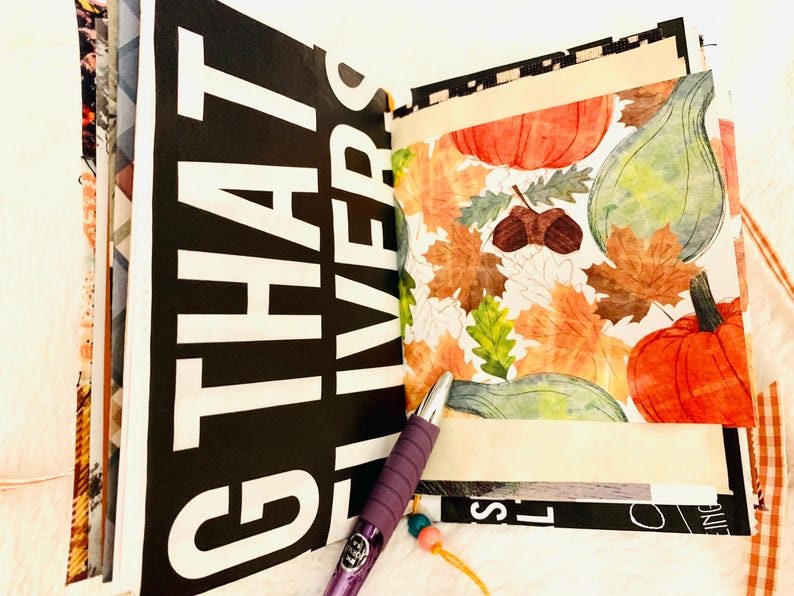 Peut inclure: Un album ouvert pr&eacute;sentant une page avec un grand texte blanc sur fond noir indiquant "THAT LIVERS" et une page adjacente d&eacute;cor&eacute;e d'un motif automnal color&eacute; de citrouilles, de courges et de feuilles d'automne. Un stylo violet est pos&eacute;.
