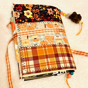 Peut inclure: Un journal recouvert de tissu avec des bandes horizontales &agrave; motifs floraux, de citrouilles et de carreaux orange. La couverture est maintenue par un ruban vichy et comporte un marque-page perl&eacute; avec une breloque ours brun sur la tranche.
