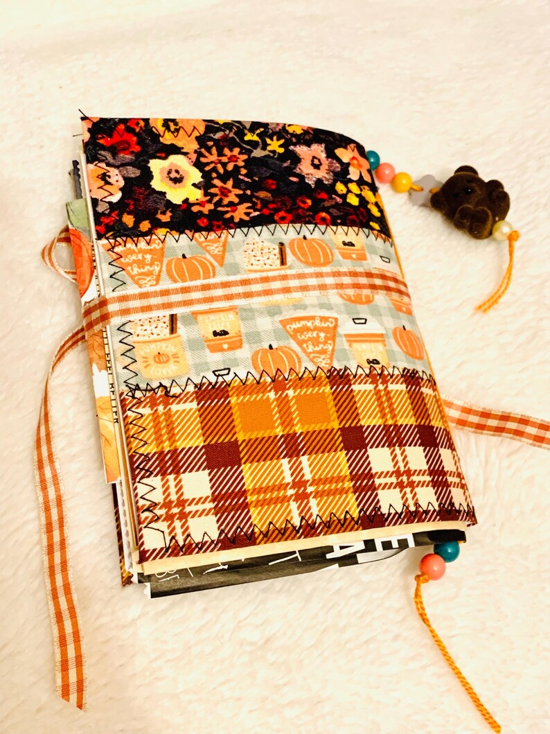 Peut inclure: Un journal recouvert de tissu avec des bandes horizontales &agrave; motifs floraux, de citrouilles et de carreaux orange. La couverture est maintenue par un ruban vichy et comporte un marque-page perl&eacute; avec une breloque ours brun sur la tranche.