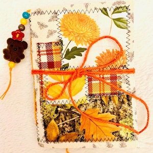 May include: A fabric-covered journal featuring a patchwork design of yellow chrysanthemums, plaid patterns, and autumn leaves. The cover is tied with orange yarn and includes a dangling charm with a brown bear and colourful beads.
