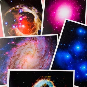 May include: A collection of five rectangular prints featuring vibrant space imagery. The prints display various celestial phenomena, including a red and gold nebula, a bright pink star cluster, a blue spiral galaxy, a blue star field, and a ring nebula.