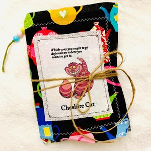 以下が含まれることがあります： 不思議の国のアリスのチェシャ猫のイラストと引用句が描かれた、布張りのノート。 ノートは紐で結ばれ、ビーズのタッセルが付いています。 生地は黒い背景にカラフルなティーポットと花の模様があります。