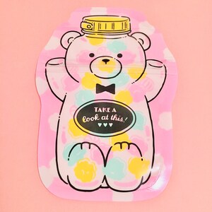 May include: Pink reusable food pouch with a cartoon bear design. The bear is wearing a bow tie and has a yellow lid on its head. The pouch has the text "Take a look at this!" on it.