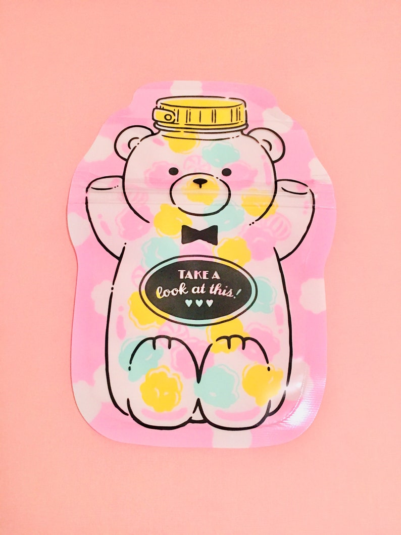 May include: Pink reusable food pouch with a cartoon bear design. The bear is wearing a bow tie and has a yellow lid on its head. The pouch has the text "Take a look at this!" on it.