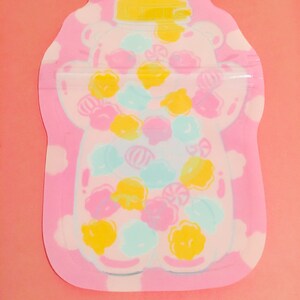 May include: A pink and white reusable food storage bag with a bear-shaped design. The bear is filled with colorful candies and has a yellow lid.