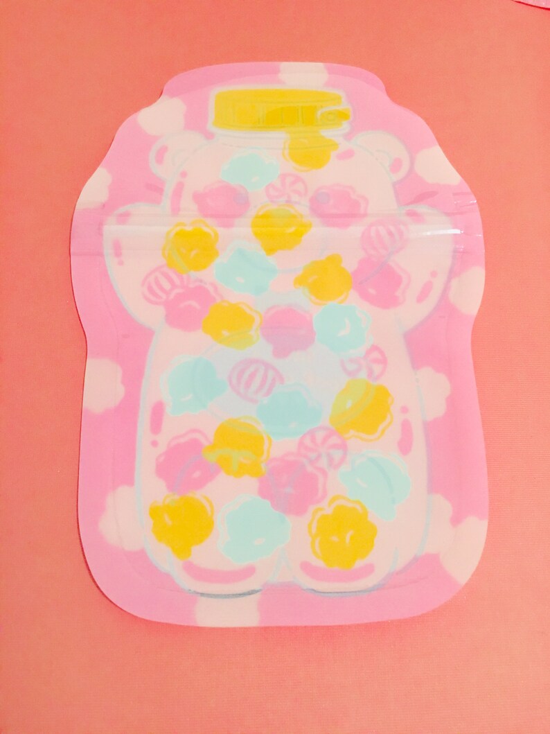 May include: A pink and white reusable food storage bag with a bear-shaped design. The bear is filled with colorful candies and has a yellow lid.