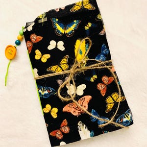 May include: A stack of black fabric items adorned with colorful butterfly patterns, tied with twine. The butterflies are in shades of yellow, orange, blue, and white. A wooden button and beads are attached to the side.