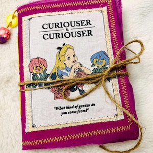 Può includere: Un diario rilegato in tessuto rosa con cuciture gialle e chiusura in spago marrone. La copertina presenta un'illustrazione di Alice nel Paese delle Meraviglie con il testo "CURIOUSER & CURIOUSER" e la citazione "Da quale tipo di giardino vieni?"