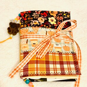 Peut inclure: Un journal recouvert de tissu compos&eacute; de trois panneaux horizontaux : un imprim&eacute; floral noir, un motif bleu clair avec des citrouilles et des tasses &agrave; caf&eacute;, et un tartan orange et jaune. Le journal est nou&eacute; avec un ruban vichy orange et un marque-page perl&eacute;.