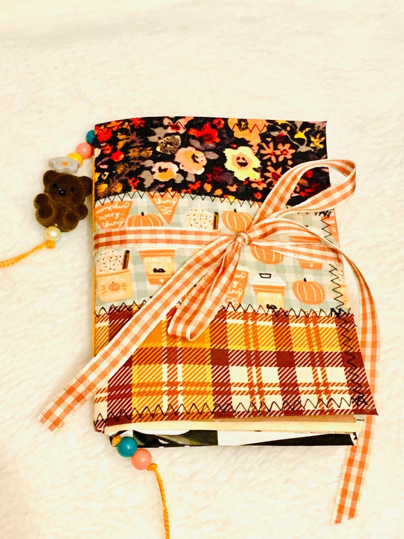 Peut inclure: Un journal recouvert de tissu compos&eacute; de trois panneaux horizontaux : un imprim&eacute; floral noir, un motif bleu clair avec des citrouilles et des tasses &agrave; caf&eacute;, et un tartan orange et jaune. Le journal est nou&eacute; avec un ruban vichy orange et un marque-page perl&eacute;.