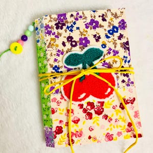 May include: A handmade fabric journal with a floral pattern in cream, purple, and red. A felt apple applique is tied with yellow cord. A beaded charm and a green spine add detail to this unique notebook.