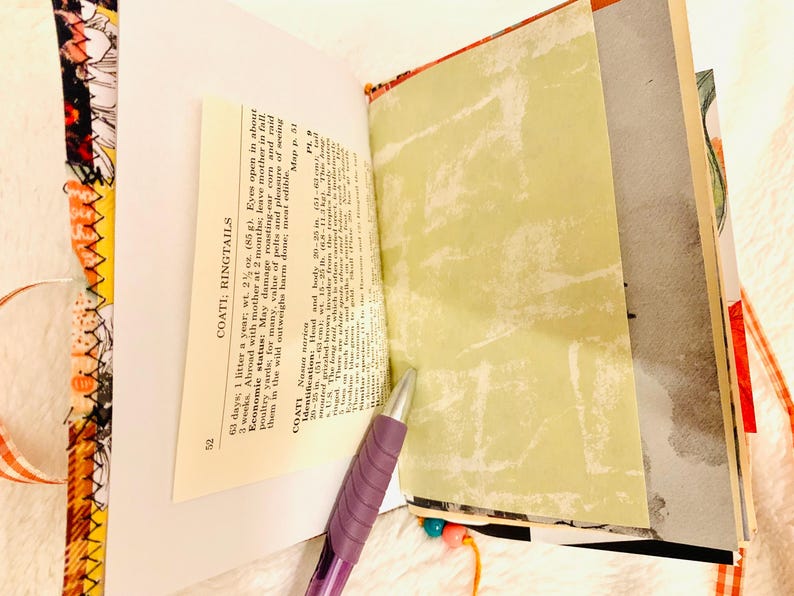Peut inclure: Un journal fait main avec une reliure en tissu aux motifs orange et noirs. Les pages ouvertes pr&eacute;sentent un texte imprim&eacute; intitul&eacute; COATI; RINGTAILS avec des d&eacute;tails incluant un poids de 85 g et une longueur de corps de 51-63 cm. Un stylo violet est pos&eacute;.