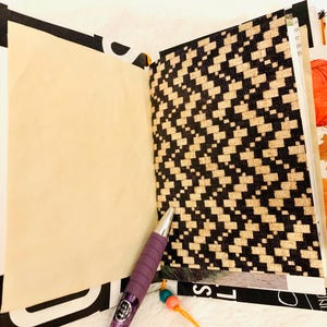 Peut inclure: Un journal ouvert avec une page vierge couleur cr&egrave;me &agrave; gauche et une page avec un motif g&eacute;om&eacute;trique noir et beige style tiss&eacute; &agrave; droite. Un stylo violet avec une prise textur&eacute;e repose en diagonale sur le bas des pages ouvertes.