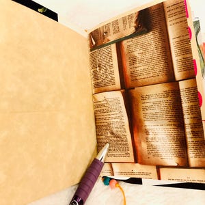 Peut inclure: Un journal ouvert pr&eacute;sentant une page vierge beige clair et une page avec un collage de texte de livre ancien. Un stylo violet &agrave; pointe argent&eacute;e repose au bas du journal. Les pages sont reli&eacute;es par des perles color&eacute;es sur un fil &agrave; la base.